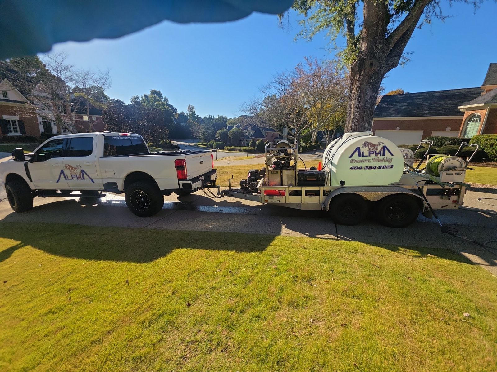 Alpha Pressure Washing pickup truck towing a black enclosed trailer lettered Alpha Pressure Washing, parked at the curb of a brick house in the evening with hoses on the trailer reels