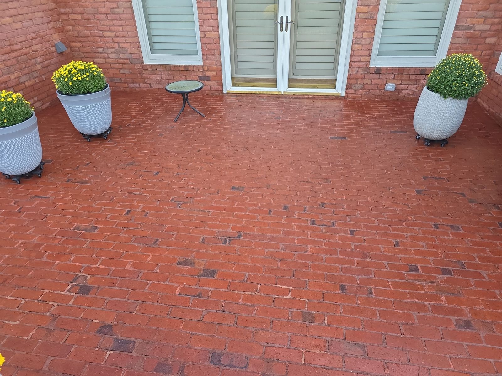 The same red brick patio in Thornberry after cleaning, with the brick color restored, a black grill, a covered table, a black metal fence, and the back of the brick house